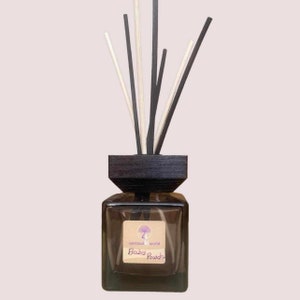 May include: A square glass diffuser bottle with a dark wooden top and reeds. The label reads "spiritual world" and "Baby Powder". The reeds are a mix of black, white, and natural wood colors. The bottle is filled with a light-colored liquid.