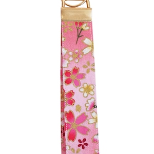 Könnte beinhalten: Rosa Schlüsselanhänger mit floralem Muster und goldfarbener Hardware. Der Stoff zeigt ein sich wiederholendes Muster aus rosa und weißen Blumen mit goldenen Akzenten. Der Schlüsselanhänger hat einen goldenen Metallring.