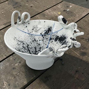 Op de afbeelding: Witte keramische kom met abstract zwart spetterontwerp en blauwe accentlijnen. De kom heeft unieke, geluste handvatten en een voetstuk. De binnen- en buitenkant zijn versierd met zwarte verfspetters.