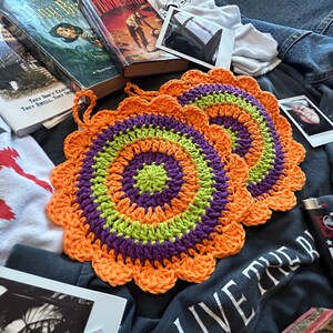 May include: Two crocheted pot holders in orange, green, and purple. The pot holders are round and have a scalloped edge.
