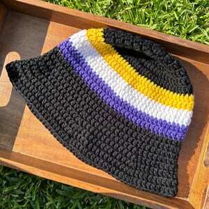 May include: A crochet bucket hat featuring a wide black base, followed by horizontal stripes of purple, white, and yellow. The hat is constructed with a textured, knotted yarn pattern throughout, creating a consistent and uniform appearance.