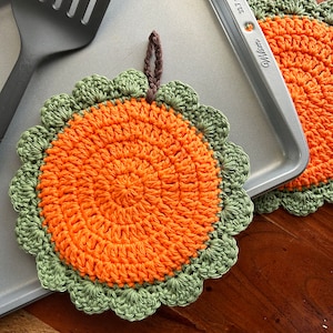 May include: Two crocheted orange and green pot holders with a scalloped edge. The pot holders are on a silver baking sheet.