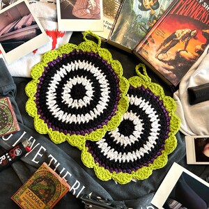 May include: Two crocheted pot holders with a black, white, and purple center surrounded by a green border. The pot holders are on a gray surface.