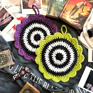 May include: Two crocheted pot holders with a black, white, and green color scheme. The pot holders have a circular design with a center circle and concentric rings. The pot holders have a scalloped edge and a loop for hanging.