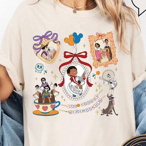 May include: A white t-shirt with a colourful graphic print featuring characters from the Disney Pixar film Coco. The graphic includes a teacup, a family portrait, and the text "We Are Family".