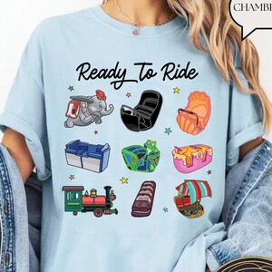 May include: Light blue t-shirt featuring the text "Ready To Ride" and colourful cartoon images of amusement park rides. The shirt is made from a soft, comfortable fabric.