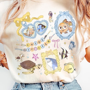 Peut inclure: Un t-shirt beige clair avec un imprimé graphique coloré de différents poissons de dessin animé, un requin, une tortue et une étoile de mer. Le texte "Guide to Fish" est imprimé sur le t-shirt.