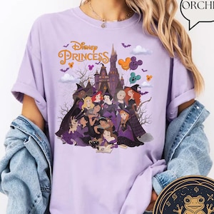May include: A light purple t-shirt featuring a graphic of Disney Princess characters dressed in witch costumes in front of a castle. The text Disney Princess is printed in gold above the characters. A speech bubble with the word ORCHID is visible.