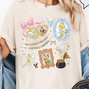 May include: Ivory t-shirt with a Tinkerbell design. The design features Tinkerbell in various poses, including flying, holding a jar of pixie dust, and standing in a heart-shaped frame. The text "Fairy Dust" is written in a whimsical font.