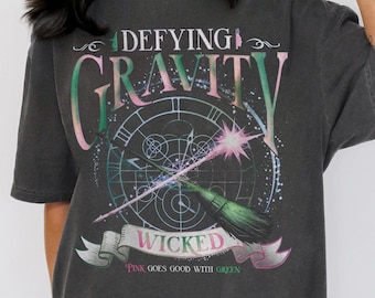 May include: A black oversized t-shirt with a graphic print of a clock, a wand, and a broom. The text on the shirt reads "Defying Gravity" and "Wicked" with the tagline "Pink goes good with green."