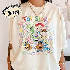 K&ouml;nnte beinhalten: Cremefarbenes T-Shirt mit einer farbenfrohen Grafik von Toy Story-Charakteren. Das Design zeigt Woody, Buzz Lightyear und andere Charaktere aus dem Animationsfilm, mit den Worten "TOY STORY" in einer verspielten Schriftart.