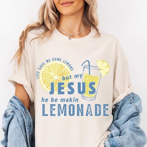 May include: Cream-colored t-shirt with a graphic of a lemon and a glass of lemonade. The text on the shirt reads "Life gave me some lemons but my Jesus he be makin lemonade" in blue and yellow.