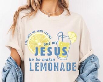 May include: Cream-colored t-shirt with a graphic of a lemon and a glass of lemonade. The text on the shirt reads "Life gave me some lemons but my Jesus he be makin lemonade" in blue and yellow.