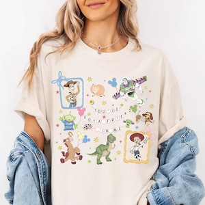Peut inclure: T-shirt crème avec un graphisme coloré de personnages de dessins animés et la phrase "You've Got a Friend in Me". Le motif comprend des images d'un cow-boy, d'un ranger de l'espace, d'un dinosaure et d'autres personnages. Le t-shirt a des manches courtes.