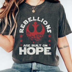 May include: Dark gray t-shirt with the text "REBELLIONS ARE BUILT ON HOPE" in white and red. The design includes a red symbol and stars. The shirt is a casual style.