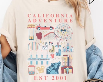 May include: Ivory-colored t-shirt with a colorful graphic of amusement park icons, including a Ferris wheel, roller coaster, and car. The text "CALIFORNIA ADVENTURE EST 2001" is printed above the graphic. A round frog design is visible.