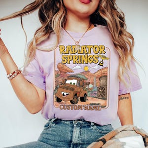 May include: A light purple t-shirt featuring a graphic of Tow Mater from the movie Cars, with the text "Radiator Springs" in yellow. The design includes a desert landscape and the words "Custom Name".