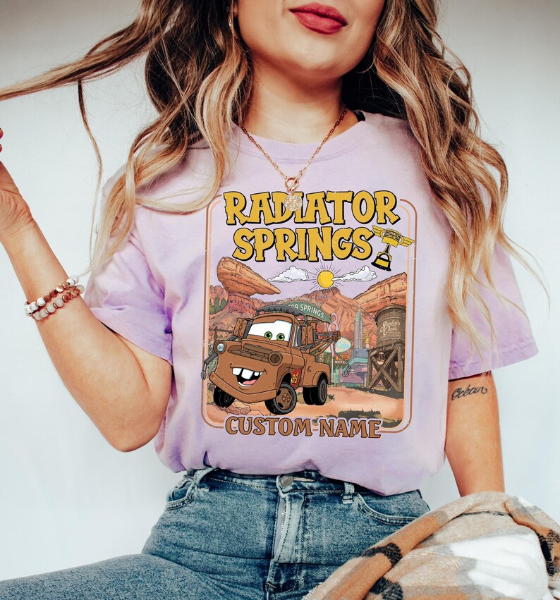 May include: A light purple t-shirt featuring a graphic of Tow Mater from the movie Cars, with the text "Radiator Springs" in yellow. The design includes a desert landscape and the words "Custom Name".