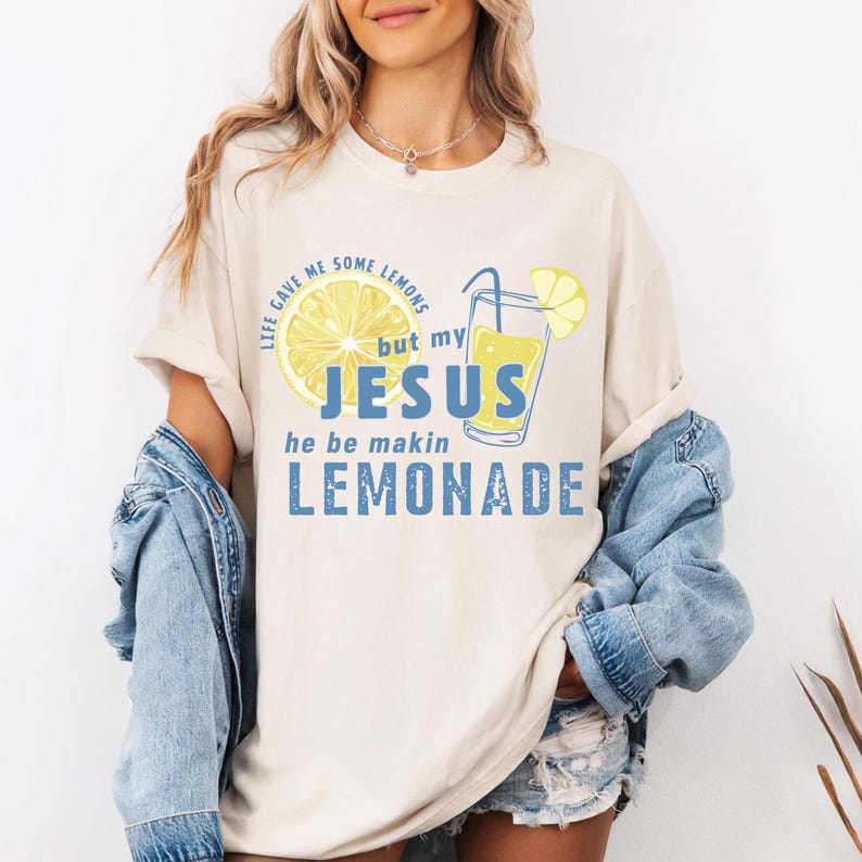 May include: Cream-colored t-shirt with a graphic of a lemon and a glass of lemonade. The text on the shirt reads "Life gave me some lemons but my Jesus he be makin lemonade" in blue and yellow.