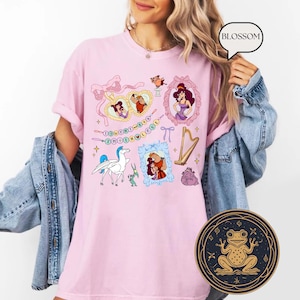 May include: A pale pink t-shirt with cartoon illustrations and the text "DON'T SAY THE LOVE". The design includes framed portraits, a harp, a pegasus, and other characters. A round sticker with a frog design is visible.