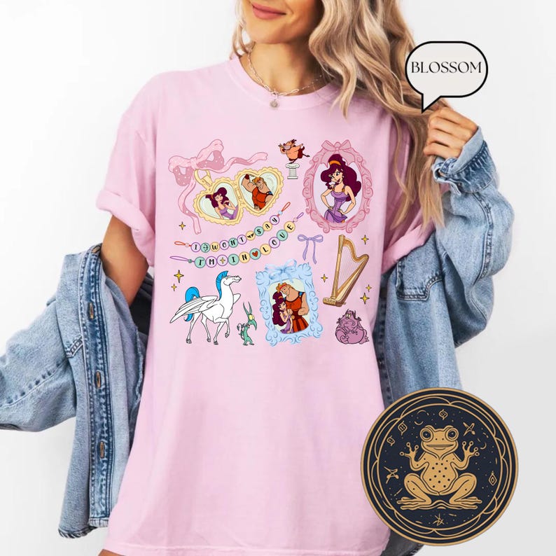 May include: A pale pink t-shirt with cartoon illustrations and the text "DON'T SAY THE LOVE". The design includes framed portraits, a harp, a pegasus, and other characters. A round sticker with a frog design is visible.