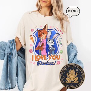 May include: Ivory-colored t-shirt featuring a cartoon graphic of a fox and a rabbit with the text "I LOVE YOU Partner!". The graphic is set within a blue shield with pink and orange accents. A round, gold-toned frog design is visible.