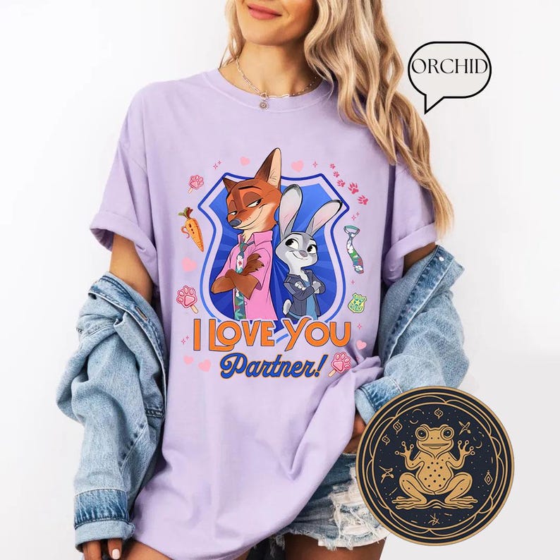 May include: A lilac-colored t-shirt featuring a cartoon image of a fox and a rabbit with the text "I LOVE YOU Partner!". The design includes a blue shield, hearts, and other small graphics. The shirt is worn with a denim jacket.
