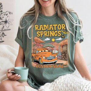 May include: Green t-shirt featuring a graphic of an orange pickup truck with the text "Radiator Springs" and "Custom Name". The design includes a desert landscape with a sun and clouds.