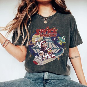 K&ouml;nnte beinhalten: Ein dunkelgraues T-Shirt mit einem Vintage-Grafikdruck von Mickey Mouse und Donald Duck, die in einem Raumschiff sitzen. Der Druck enth&auml;lt den Text "Space Mountain".
