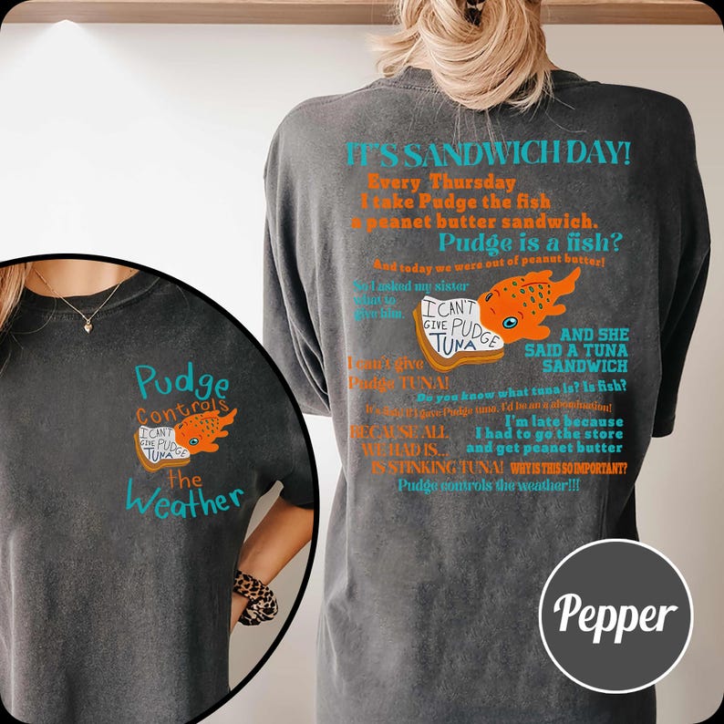 Puede incluir: Camiseta gris oscuro con un gr&aacute;fico de un pez naranja y el texto "Pudge Controls The Weather". La parte trasera de la camiseta tiene un dise&ntilde;o humor&iacute;stico con tem&aacute;tica de s&aacute;ndwich con texto y una ilustraci&oacute;n de pez. La camiseta es de Pepper.