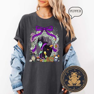 Op de afbeelding: Donkergrijs t-shirt met een cartoon illustratie van een groenhuidig personage en een zwarte kraai, omringd door bloemen, pompoenen en schedels. Een paarse strik en het woord "PEPPER" zijn ook zichtbaar.
