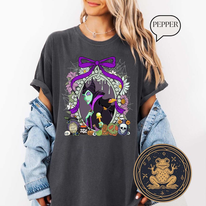 Op de afbeelding: Donkergrijs t-shirt met een cartoon illustratie van een groenhuidig personage en een zwarte kraai, omringd door bloemen, pompoenen en schedels. Een paarse strik en het woord "PEPPER" zijn ook zichtbaar.