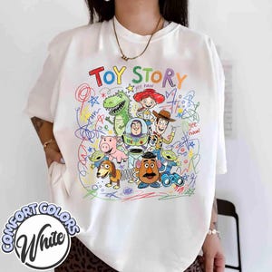K&ouml;nnte beinhalten: Wei&szlig;es Comfort Colors T-Shirt mit einem farbenfrohen Toy Story-Charakter-Design. Das Design zeigt Woody, Buzz Lightyear, Jessie, Rex und andere Charaktere mit dem Schriftzug "Toy Story". Das T-Shirt hat eine lockere Passform.