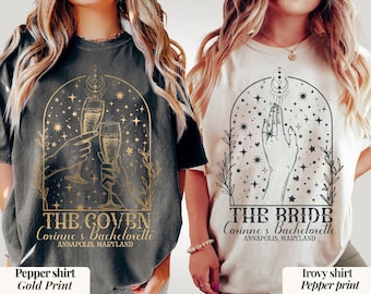 May include: Two t-shirts, one pepper-colored and one ivory-colored, with graphic designs. The pepper shirt has a gold print of two hands toasting champagne glasses, with text "THE COVEN" and "Corinne's Bachelorette, Annapolis, Maryland." The ivory shirt has a pepper print of a hand reaching up, with text "THE BRIDE" and "Corinne's Bachelorette, Annapolis, Maryland."
