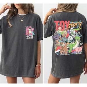 May include: Dark gray t-shirt featuring a colorful graphic from the movie Toy Story. The front of the shirt has a pink bear graphic with the word "Name". The back of the shirt has a Toy Story graphic with the words "Toy Story" and "Since 1995".