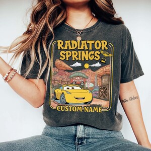 May include: Dark gray t-shirt featuring a yellow cartoon car with the text "RADIATOR SPRINGS" and "CUSTOM-NAME". The design includes a desert landscape and the words "RACERS". The shirt is a comfortable, casual wear item.