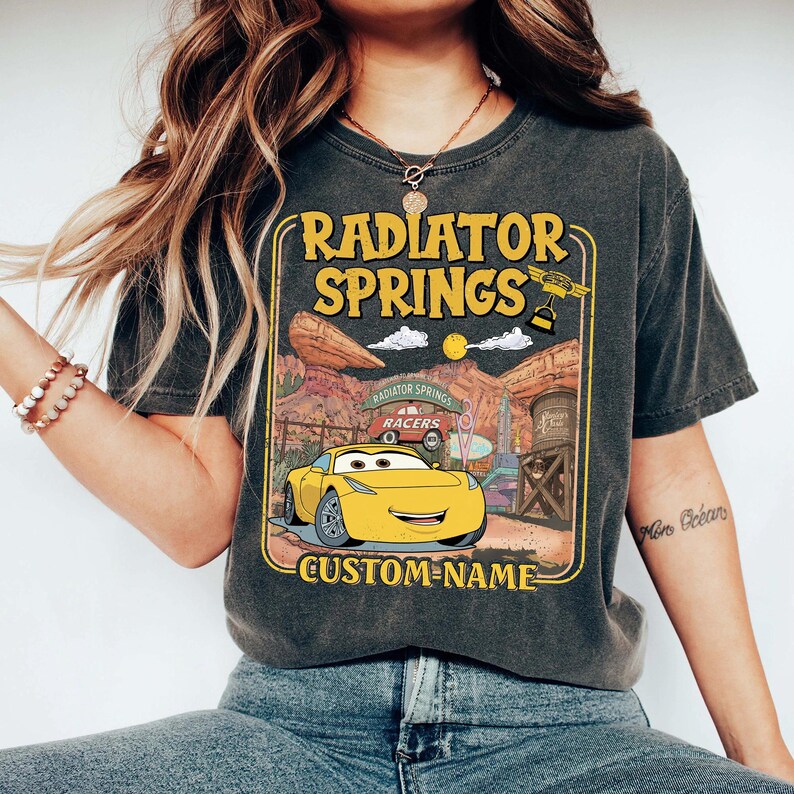 May include: Dark gray t-shirt featuring a yellow cartoon car with the text "RADIATOR SPRINGS" and "CUSTOM-NAME". The design includes a desert landscape and the words "RACERS". The shirt is a comfortable, casual wear item.