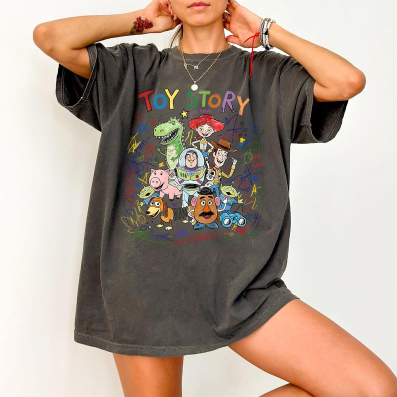 K&ouml;nnte beinhalten: Dunkelgraues T-Shirt mit dem Schriftzug "TOY STORY" in bunten Buchstaben &uuml;ber einer Grafik von Zeichentrickfiguren. Das Design umfasst Charaktere wie Woody, Buzz Lightyear und Rex. Das Shirt hat einen Vintage-Look.
