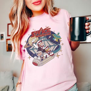 K&ouml;nnte beinhalten: Rosafarbenes T-Shirt mit einem Vintage-Grafikdruck von Mickey Mouse, Donald Duck und Goofy, die auf der Space Mountain fahren. Die Grafik enth&auml;lt den Text "Space Mountain".