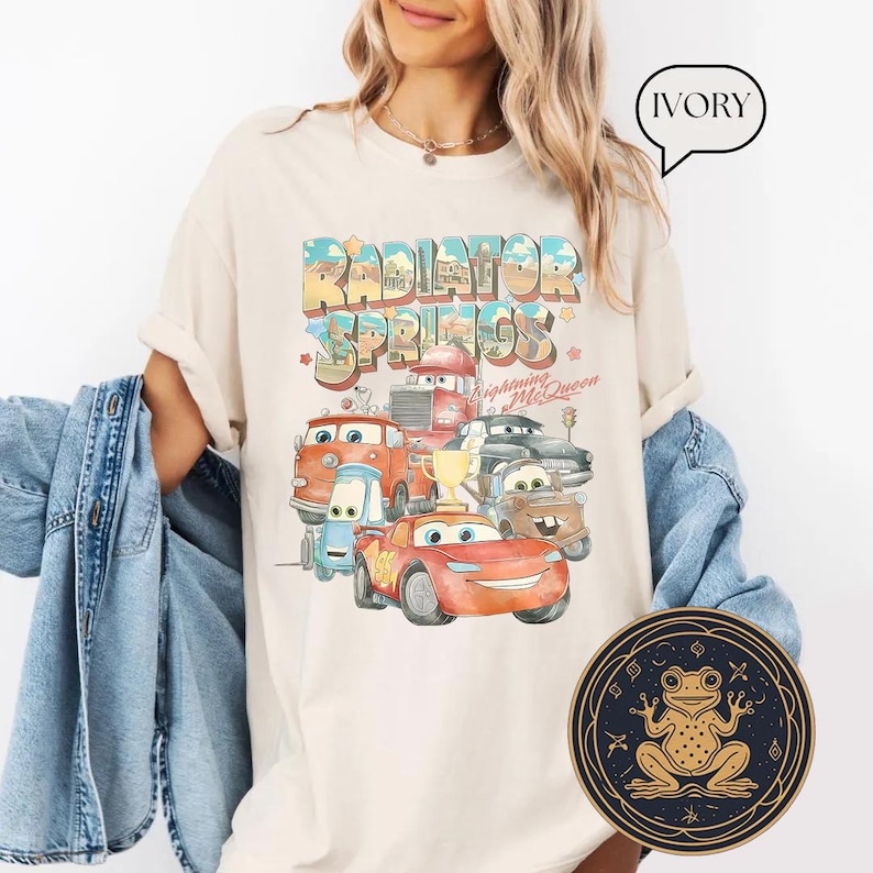May include: A white t-shirt with a vintage-style graphic of the Cars characters from the movie Cars. The graphic features the text "Radiator Springs" and includes Lightning McQueen, Mater, and other characters. The shirt is available in ivory.