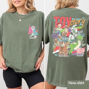 May include: Moss green t-shirt featuring a Toy Story design. The front has a cartoon character and the word "Name". The back displays the Toy Story logo and characters including Woody, Buzz, and Rex. The shirt is a relaxed fit.