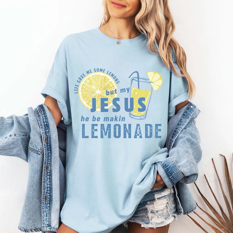 May include: Light blue t-shirt with a graphic of a lemon slice, a glass of lemonade, and the text "Life gave me some lemons but my Jesus he be makin lemonade" in blue. The shirt has short sleeves.