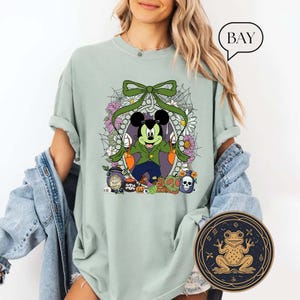 Op de afbeelding: Lichtgroen T-shirt met een Halloween-ontwerp met Mickey Mouse verkleed als vampier, omringd door bloemen en spinnenwebben. Het ontwerp bevat pompoenen, schedels en drankflessen. Een ronde kikkerafbeelding is rechtsonder zichtbaar.