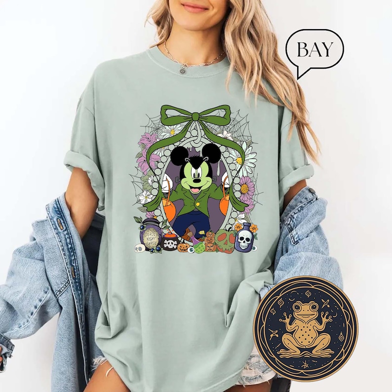 Op de afbeelding: Lichtgroen T-shirt met een Halloween-ontwerp met Mickey Mouse verkleed als vampier, omringd door bloemen en spinnenwebben. Het ontwerp bevat pompoenen, schedels en drankflessen. Een ronde kikkerafbeelding is rechtsonder zichtbaar.