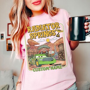 May include: Pink t-shirt featuring a graphic of Radiator Springs from the movie Cars. The design includes a green race car, the text "Radiator Springs," and a space for a custom name. The person is holding a black mug.