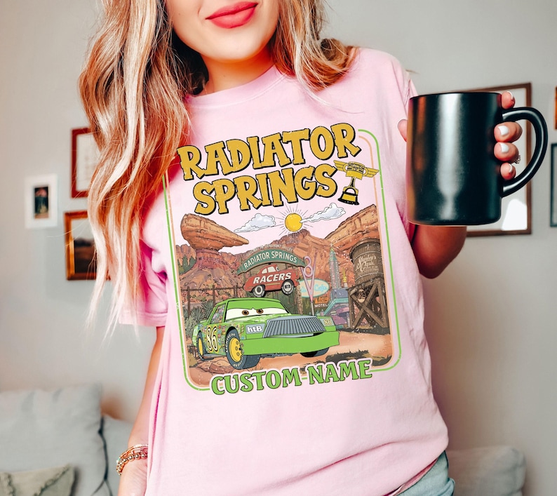 May include: Pink t-shirt featuring a graphic of Radiator Springs from the movie Cars. The design includes a green race car, the text "Radiator Springs," and a space for a custom name. The person is holding a black mug.