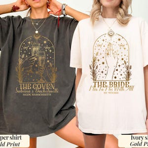 May include: Two short-sleeve t-shirts, one pepper gray and one ivory, each featuring a gold-colored graphic print. The pepper shirt displays two hands holding champagne glasses with the text THE COVEN, Sabrina's Bachelorette, and SALEM, MASSACHUSETTS. The ivory shirt shows a hand with the text THE BRIDE, Miss To Mrs With All MY WITCHES, and EST 2026.