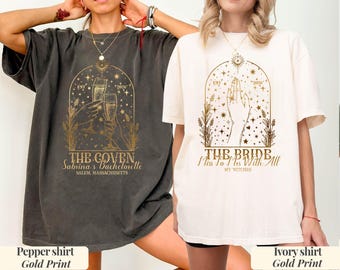 Pode incluir: Duas t-shirts de manga curta, uma cinza pimenta e outra marfim, cada uma com uma impressão gráfica dourada. A t-shirt cinza mostra duas mãos segurando taças de champanhe com o texto THE COVEN, Sabrina's Bachelorette e SALEM, MASSACHUSETTS. A t-shirt marfim mostra uma mão com o texto THE BRIDE, Miss To Mrs With All MY WITCHES e EST 2026.