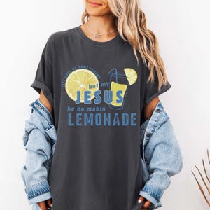 May include: Dark grey t-shirt with a graphic design featuring a lemon and a glass of lemonade. The text reads "Life gave me some lemons but my Jesus he be makin lemonade" in blue and yellow.