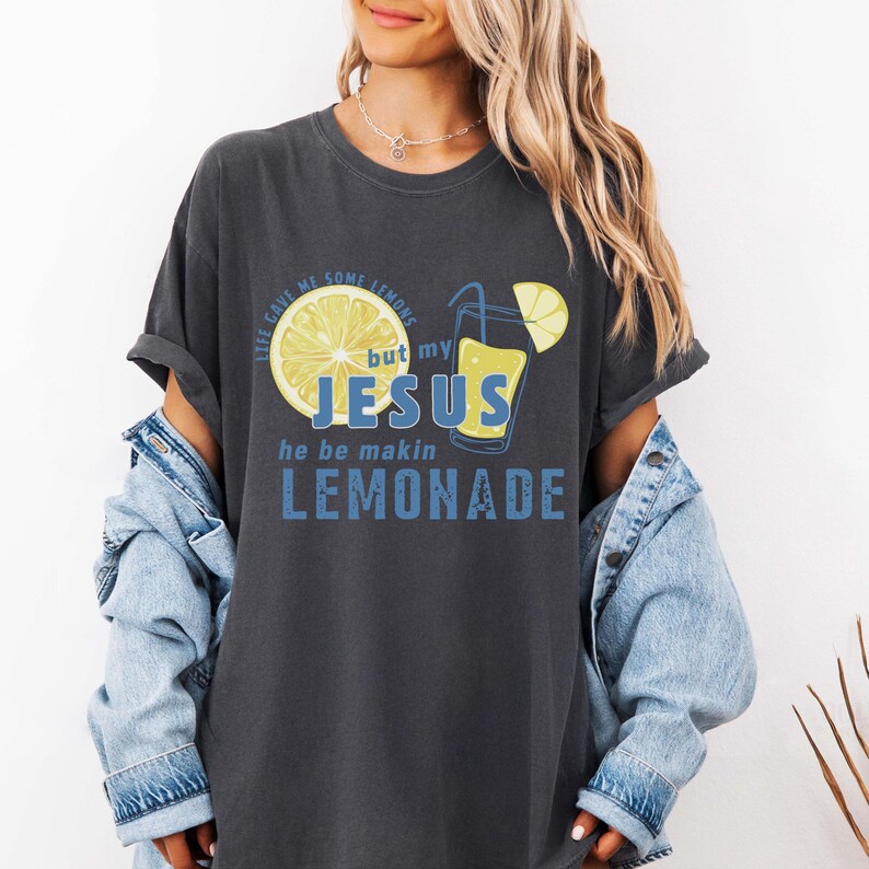 May include: Dark grey t-shirt with a graphic design featuring a lemon and a glass of lemonade. The text reads "Life gave me some lemons but my Jesus he be makin lemonade" in blue and yellow.